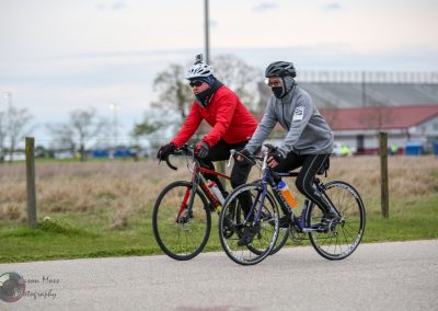 Cyclists riding on a paved trail during daytime, wearing helmets, masks, and warm sportswear.