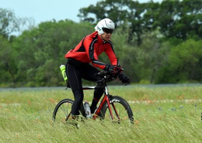 Bright cyclist riding mountain bike on grassy trail with lush green trees in the background.