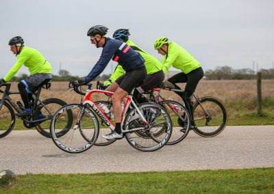 Bike riders in bright neon and athletic gear on a scenic outdoor trail with open fields.