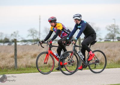 Two cyclists riding bikes outdoors on a paved path, wearing helmets and sports gear for biking.