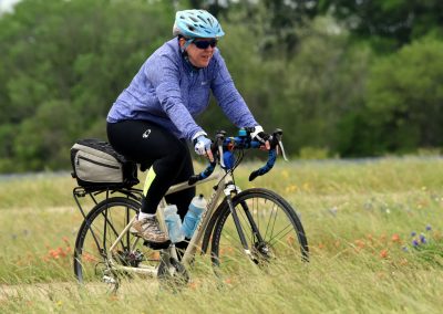 Mid-adult cyclist riding a bike through a grassy field with green trees in the background.