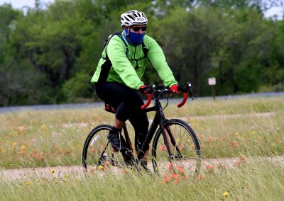 Vibrant cyclist riding through grassy field on Bluebonnet Express trail with colorful wildflowers and lush green trees in background.
