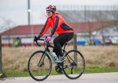 Girl riding a black road bike on a paved trail, wearing a red and black cycling jacket and helmet.