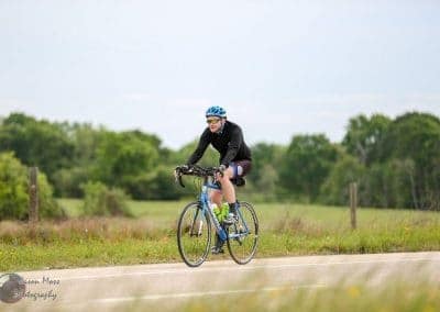Bright blue bicycle on scenic rural bike trail with rider wearing helmet and sunglasses, promoting Bluebonnet Express cycling event.