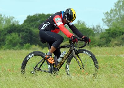 High-performance cyclist riding a black and yellow Trek Madone bike through grassy fields in outdoor sports setting.