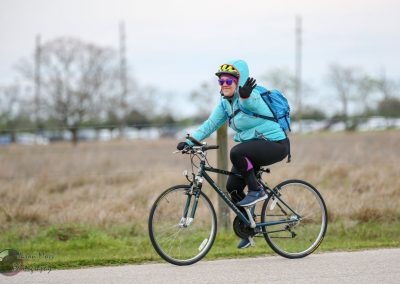 Cyclist waving while biking on a scenic trail, wearing a Bluebonnet Express jacket and gear.