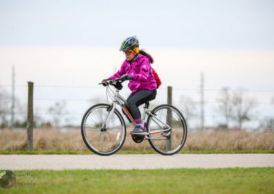 Vibrant girl cycling outdoors with a pink jacket, helmet, and backpack on a gray bike in a scenic park setting.
