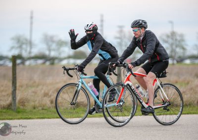 Wearing a helmet and sunglasses, two cyclists ride together outdoors on a paved trail.