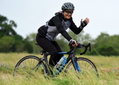 Bright woman riding a Bluebonnet Express bike through green fields, smiling and waving, enjoying outdoor cycling.