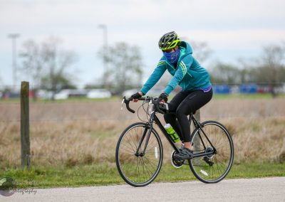 Bright blue cycling jacket, helmet, and face mask on a woman riding a black road bike outdoors.