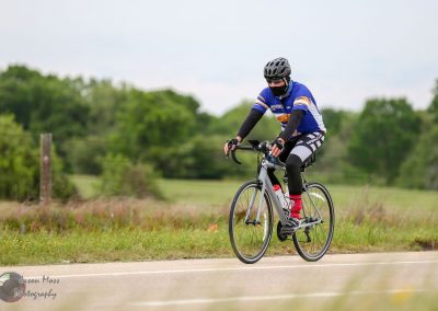Bright blue cyclist jersey with black biking gear riding on bike trail with lush green background.