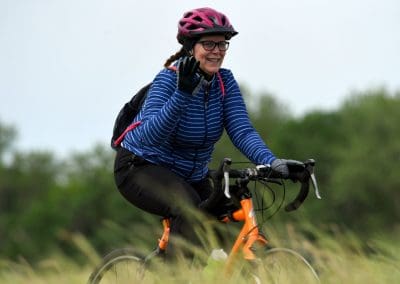 Bright woman cycling outdoor in windbreaker, wearing pink helmet, glasses, and smiling.
