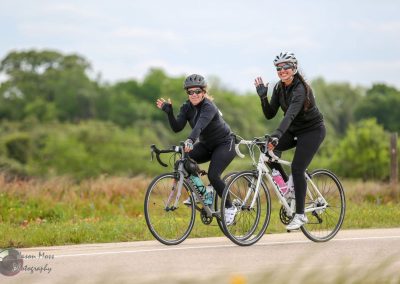 Two women riding bikes outdoors, smiling and waving, on a scenic bike trail with lush greenery.