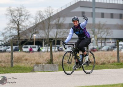 Bright female cyclist waving on a scenic bike trail with modern building in the background.
