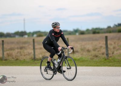 1. Cyclist riding a black road bike on a rural trail with open fields in the background.