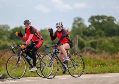 Bright female cyclists riding on a scenic countryside road, promoting Bluebonnet Express bike tours.