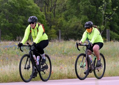 Vibrant cyclists riding on scenic trail amidst wildflowers and lush greenery, promoting outdoor biking adventures at Bluebonnet Express.
