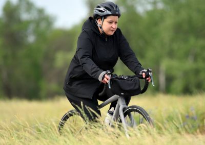 Young woman riding a bicycle on grassy trail, wearing helmet and black jacket, enjoying outdoor cycling.