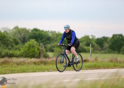 Bright woman cycling outdoors on a paved rural road with greenery, wearing a helmet and athletic attire.