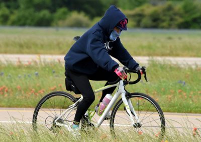 Bright cyclist wearing a hoodie, face mask, and gloves riding a bike through a scenic field.