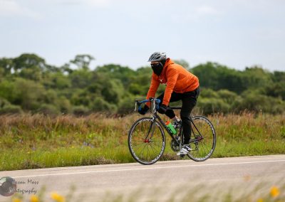 Bright orange cycling jacket with black pants and helmet riding a bike through a scenic rural landscape, showcasing Bluebonnet Express cycling route.