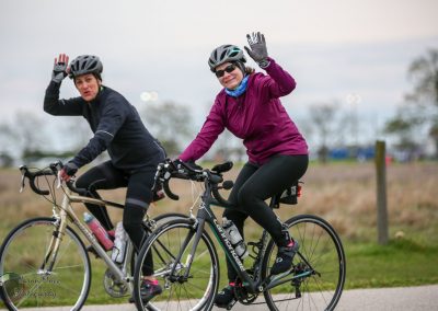 Women riding bicycles, smiling and waving, in a scenic outdoor setting for Bluebonnet Express event.