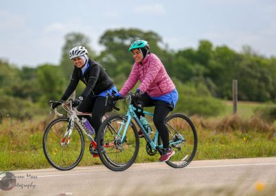 Brightly dressed women cycling on scenic rural trail during daytime, promoting Bluebonnet Express bike tours.