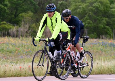 Brightly dressed cyclists riding on a scenic trail with wildflowers and green foliage in the background.