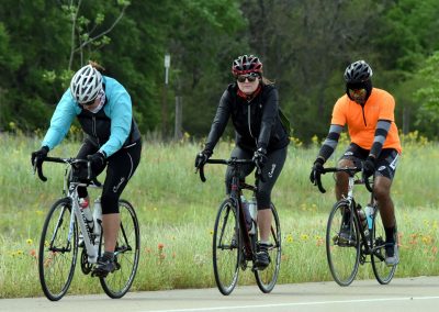Brightly dressed cyclists riding on a scenic bike trail with wildflowers and greenery in the background.