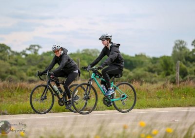 Two women cycling on scenic rural trail with lush greenery and overcast sky, enjoying outdoor biking adventure.