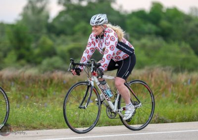 Elegant female cyclist riding a road bike outdoors with greenery in the background for Bluebonnet Express event.