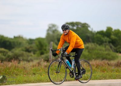 Vibrant cyclist wearing orange jacket riding a black road bike on a rural trail with lush greenery.