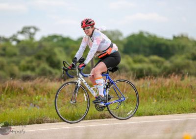 Bright female cyclist riding a blue and white road bike on a scenic countryside trail during daytime.