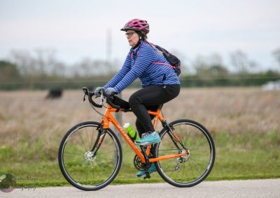 Bright orange hybrid bike with a woman cyclist wearing a pink helmet, blue striped jacket, and black pants riding on a paved trail.
