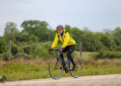 Bright yellow cycling jacket on male cyclist riding on rural road with lush greenery, promoting Bluebonnet Express bike tours.