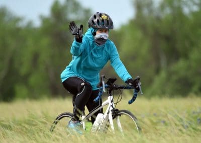 Woman cyclist wearing a helmet and face mask, riding a bike outdoors in a grassy field.