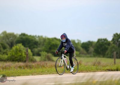 Bright woman cycling outdoors in a hoodie, wearing a mask and helmet, on a scenic rural road, promoting Bluebonnet Express bike services.