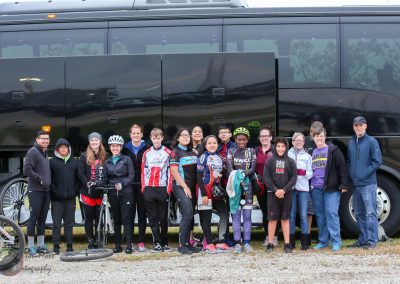 Group of young cyclists and their support team standing in front of a large black bus, promoting Bluebonnet Express cycling trips.