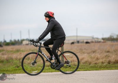 Bike commuter riding a black and red Trek mountain bike on a paved trail in a rural area during overcast weather.