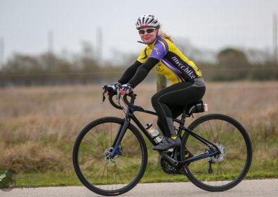 Cyclist riding a bicycle outdoors with Bluebonnet Express branded jersey, on a scenic trail.