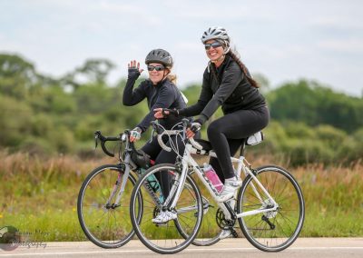 Bright women smiling while riding bicycles on the scenic Bluebonnet Express trail on a clear day.