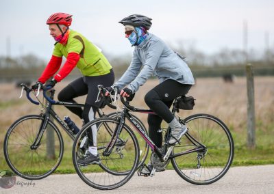 Smiling cyclists riding on a scenic rural trail in cool weather, wearing helmets and cycling gear for outdoor adventure.