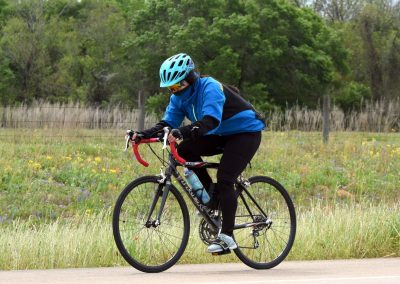 Bright blue helmet and jacket, cyclist riding on scenic road with wildflowers and green trees.