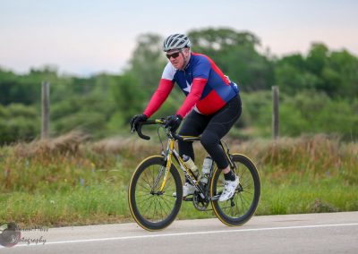 Vibrant cyclist in Bluebonnet Express gear riding scenic rural road during daytime.