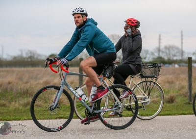 Riders enjoying a scenic bike ride on a paved trail with a cyclist in the foreground and a woman behind, both wearing helmets.