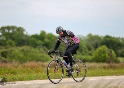 Vegan cyclist riding on a rural road, raising hand in greeting, surrounded by lush green landscape.