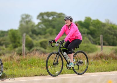 Bright pink jacket woman riding a bicycle on a scenic outdoor trail during daytime for Bluebonnet Express tour, joyful cycling activity.