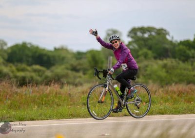 Bright female cyclist riding a bike on a scenic outdoor trail, smiling and waving during a sunny day.