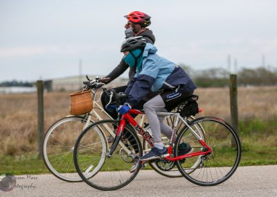 Two cyclists riding bikes on a paved path in an open, rural area during daytime, dressed in outdoor cycling gear.