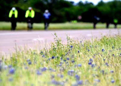 Blooming wildflowers on roadside in Texas with cyclists in the background, celebrating Bluebonnet Express bike tour.